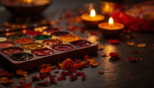 An open paint set in golds, reds and jewel tones beside glowing diya oil lamps and scattered marigold petals — a Diwali craft table.